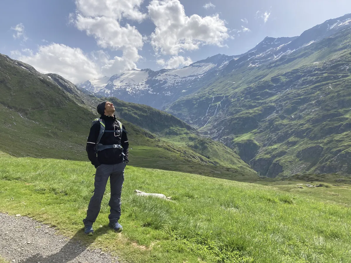 Marcel Schreyer auf einer Bergwiese, den Blick nach oben zum Himmel, dahinter ein Tal und schneebedeckte Gipfel