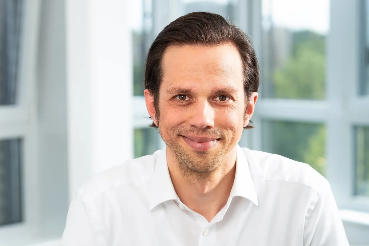 Marcel Schreyer im weißen Hemd, lächelnd, vor einer Fensterfront mit Bäumen dahinter